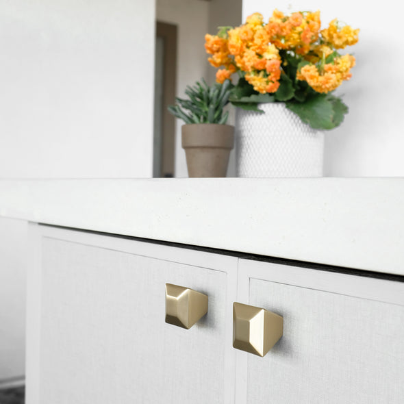 Two Satin Brass Diamond knobs installed on tan cabinet doors with orange flowers and a succulent on the countertop
