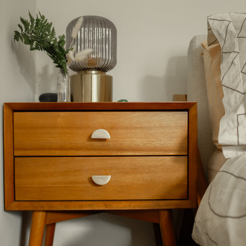 Two satin brass half moon knobs on an end table in a bedroom with white linen and white walls. Mid century modern style.