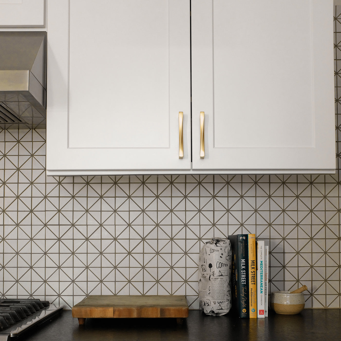 Solid Brass Twist Cabinet Pulls in a White Kitchen with Cookooks and a Cutting Board and a geometric backsplash. 