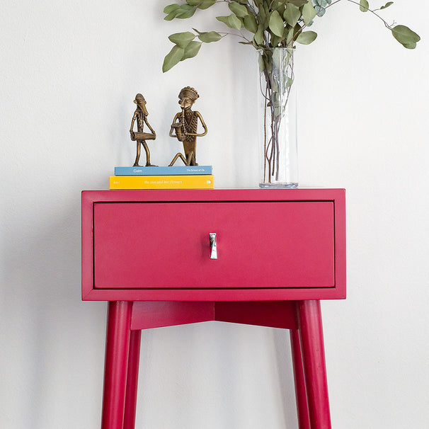 Red side table with a drawer, books, and decorative items on a white background with polished chrome knob.