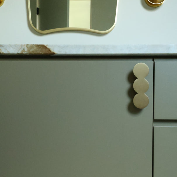 Close-up of a light gray cabinet with a round handle on a marble countertop finished in solid brass.
