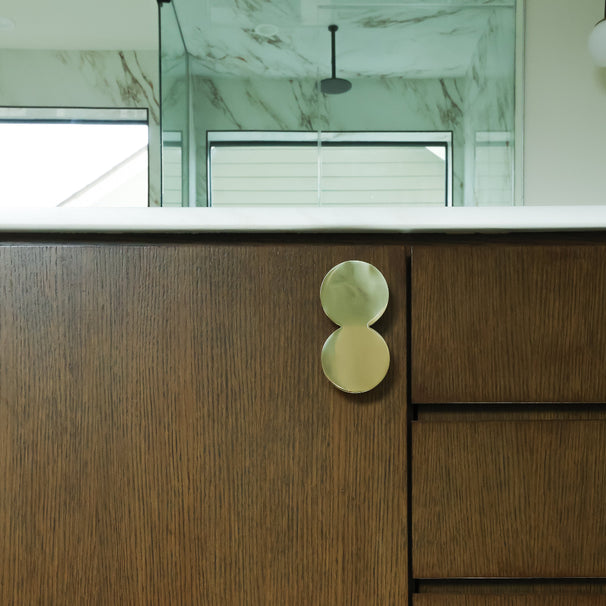 A close-up of a modern bathroom vanity featuring a dark wood cabinet with a distinctive double-circle brass pull handle and a white marble countertop.