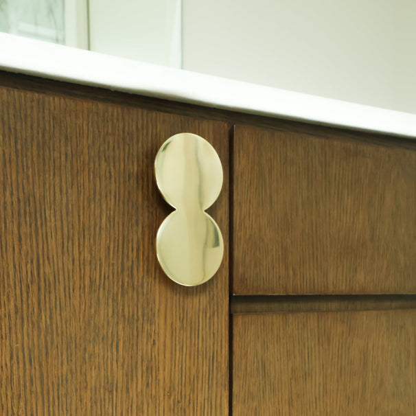 A close-up of a polished gold double-oval cabinet pull handle mounted on a dark brown wood cabinet door.