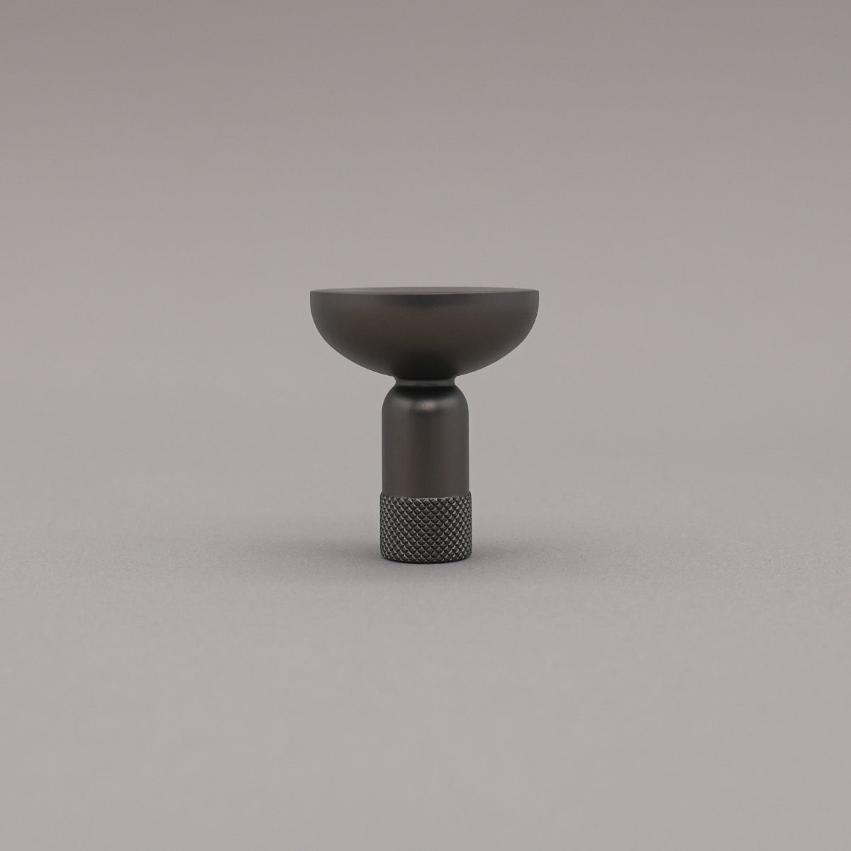 Upright view of skinny knurled knob in graphite finish on grey background.