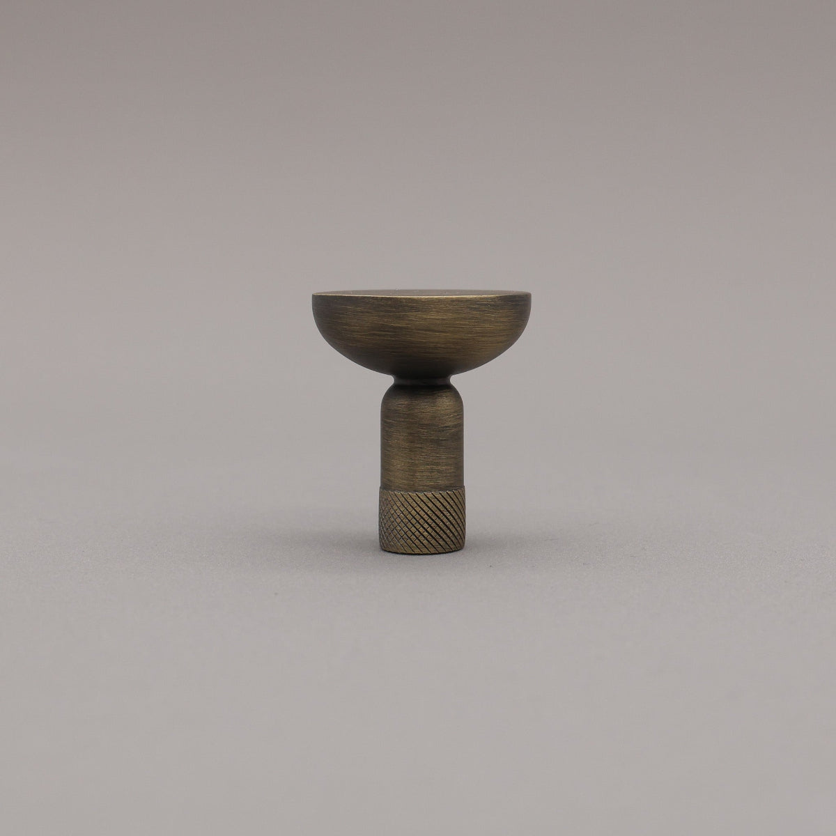 Upright view of skinny knurled knob in aged brass finish on grey background.