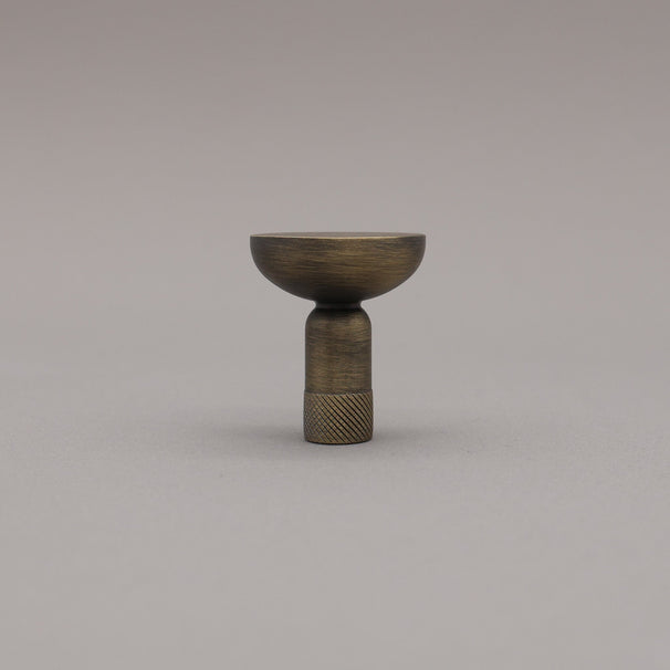 Upright view of skinny knurled knob in aged brass finish on grey background.