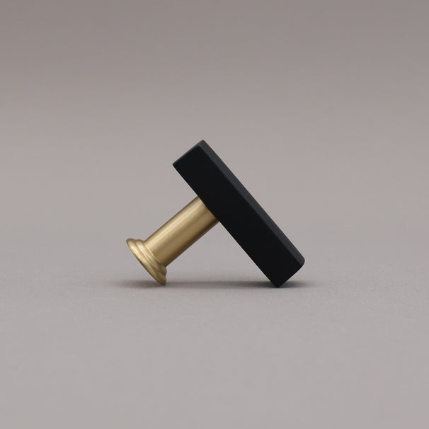 Angled satin brass and black t-knob brass cabinet knob on gray background.