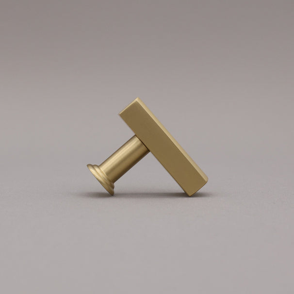Leaning satin brass t-knob brass cabinet knob on gray background.