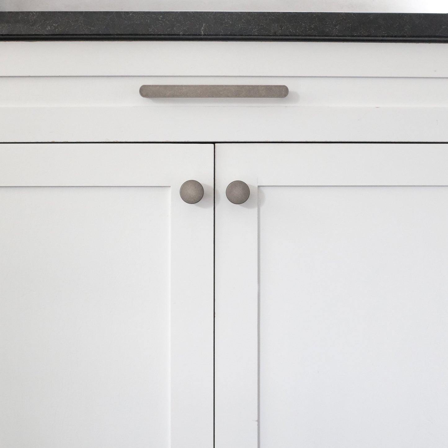 Ribbed knobs and pull on white kitchen cabinets with black counter.