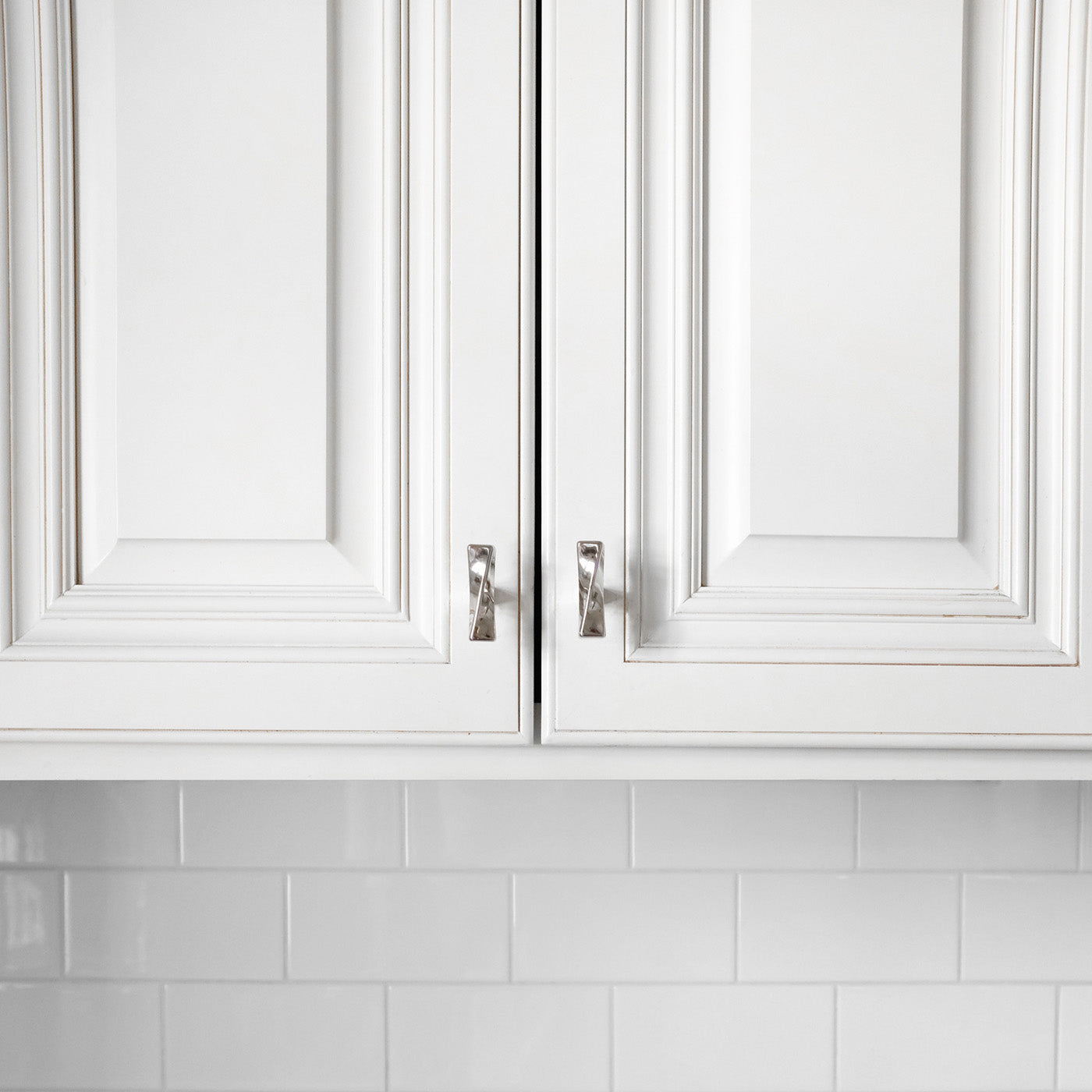 Close-up of white kitchen cabinets with polished nickel knobs against a white tiled backsplash.