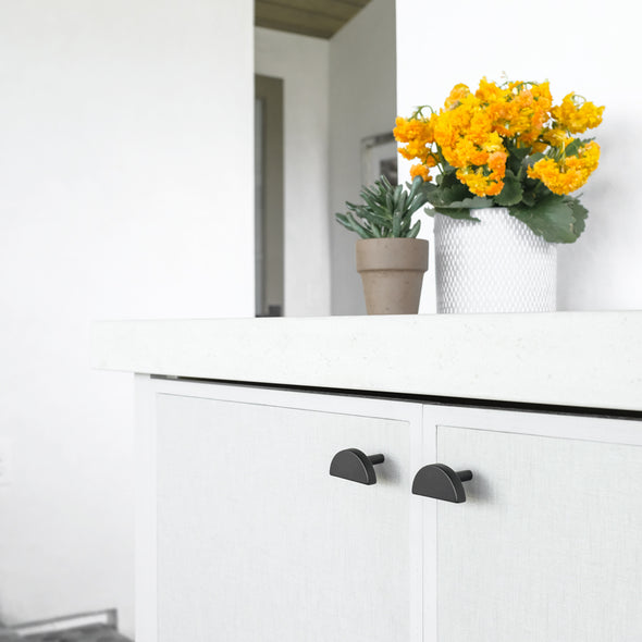 Two Hapny Matte Black Half Moon cabinet knobs installed on white cabinet drawers with orange flowers on the countertop