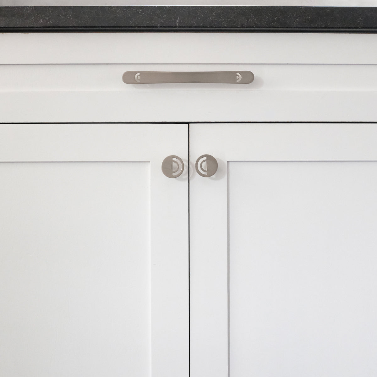 One 8" Hapny Horizon cabinet pull and two Horizon knobs in Satin Nickel installed on white cabinet doors and drawers