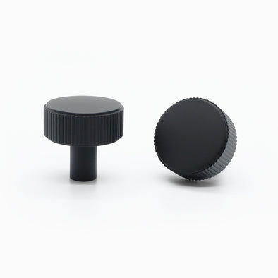 Two Matte Black Hapny Ribbed knobs, one standing and one front-facing while laying down