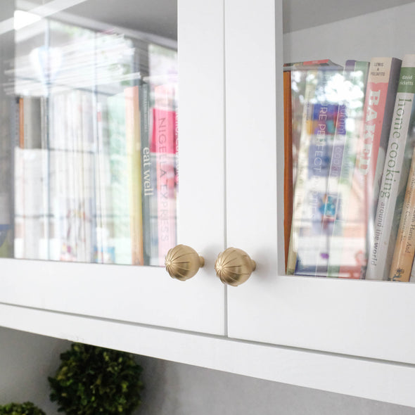 Two Satin Brass Hapny Sunburst knobs installed on white cabinet doors with glass panels filled with cookbooks