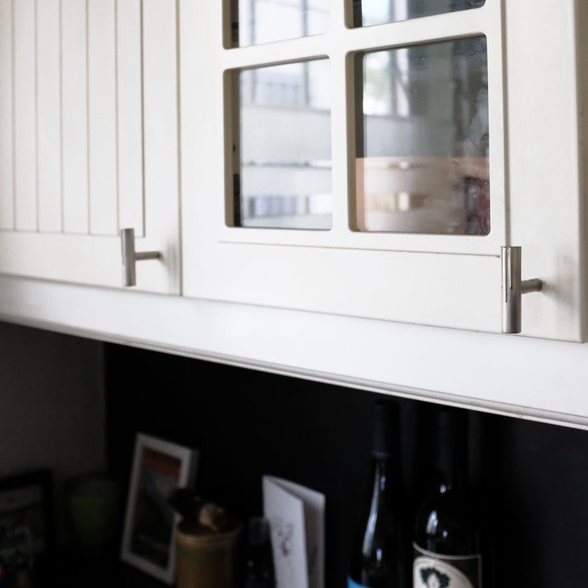 Two Hapny Sunburst t-knobs in a Satin Nickel finish installed on white kitchen cabinet doors with glass panels