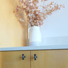 Two Matte Black Hapny Twist t-knobs installed on brown cabinet doors with a vase of pink leaves on the countertop
